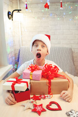 A child in a Santa Claus hat is hugging boxes of Christmas gifts in surprise with his mouth open. Made by hand. Bright interior. Vertical orientation.