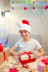 A smiling boy wraps Christmas presents in a Santa Claus hat. Christmas gifts with your own hands. Home leisure with children in the New Year.