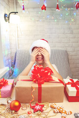 A child in a Santa Claus hat has closed his eyes with his hands and is waiting for a New Year's gift. A large box with a red bow stands in front of the boy.