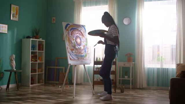 Young black woman student has fun at home dancing and painting on big canvas, holding brush and palette with paints, transmits inner creative energy into her work, feeling free and happy, Slow motion.