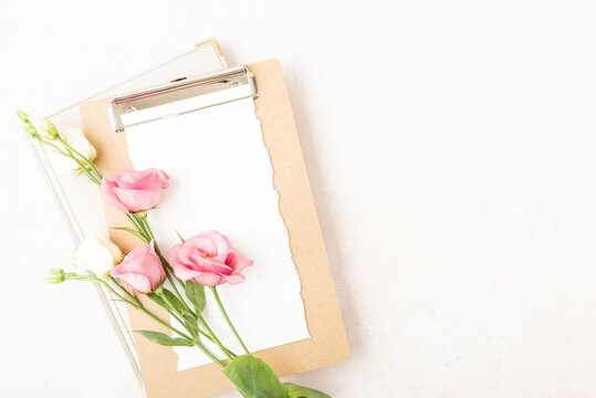Blank paper clipboard, rose and white blooming flowers. Flat lay, top view with copy space. Home office or table desk in rose, white, gold colors. Mockup for wedding invitation or menu background