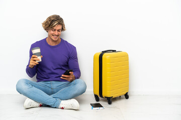 Young blonde man with suitcase sitting on the floor holding coffee to take away and a mobile