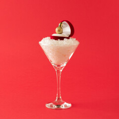 Cocktail glass with snow and red ring gift box with gold Christmas ball. Creative New Year celebration concept.