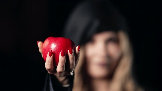 Woman as witch with an evil grin laugh in black offers red apple as symbol of temptation, poison. Fairy tale white snow concept, halloween, cosplay.