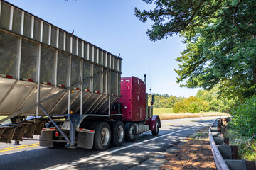 Fototapeta na wymiar Purple big rig classic semi truck transporting cargo in open bulk semi trailer running on the narrow road with protection fence