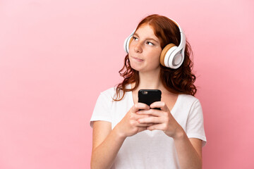 Teenager reddish woman isolated on pink background listening music with a mobile and thinking