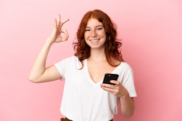 Teenager reddish woman isolated on pink background using mobile phone and doing OK sign