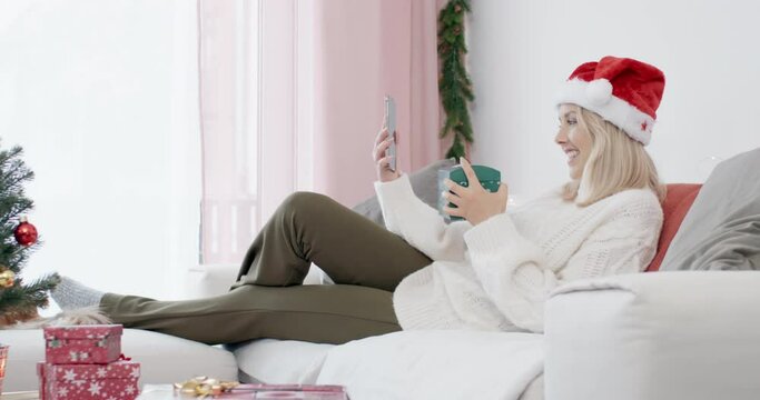 Young smiling woman wearing a santa hat making video call on cosy couch and opens christmas gift in front of selfi cam shot in 4k