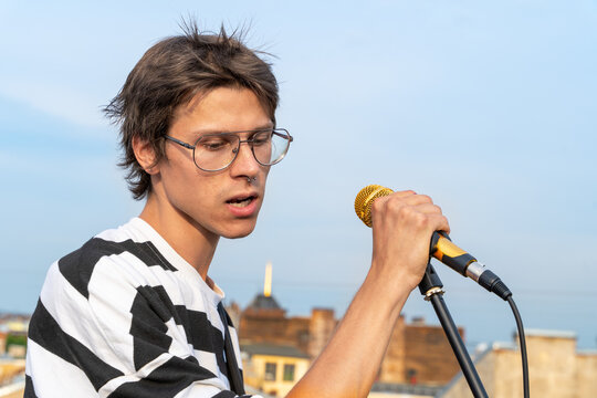 Portrait of young guy with glasses, singing with gold microphone on roof, his hair is disheveled by wind. Romantic image of singer during musical outdoor concert or reading poetry by poet.