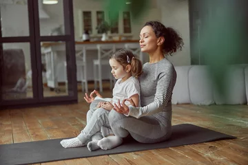 Fototapeten Relaxed woman is sitting with little girl in lotus position and doing meditation at home © Yaroslav Astakhov