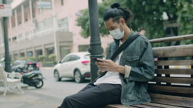 Asian man wearing a mask and using a smartphone checking social media while sitting on a bench beside the street in the city.