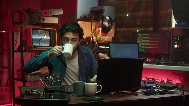 Hacker using laptop, drinking coffee. Young Spanish man developing program in dark room. Professional programmer infecting system with cyber virus, typing on computer keyboard, breaking password.