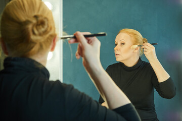 Mature woman in a black turtleneck does make-up, applies blush with a cosmetic brush, looking in the mirror