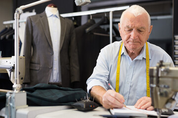 Elderly man tailor uses notepad to record jacket measurements in workshop