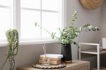 Vase with eucalyptus, books and reed diffuser on shelf near window - obrazy, fototapety, plakaty