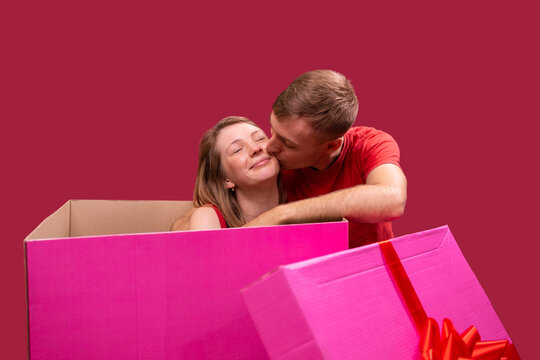 Close up photo of a loving couple, girlfriend sitting inside the big present box of pink color with closed eyes in pleasure of her boyfriend kiss on the cheek.