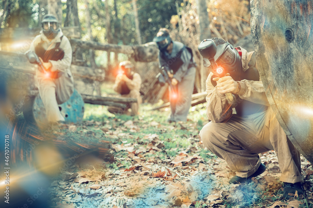 Poster Team of male and female paintball players with marker guns aiming on gamefield outdoors