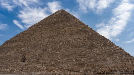 Fototapeta na wymiar The Great Pyramid of Cheops on the background of a blue sky with picturesque clouds. On the wall, made of ancient boulders, you can see the entrance inside. Egypt. Giza