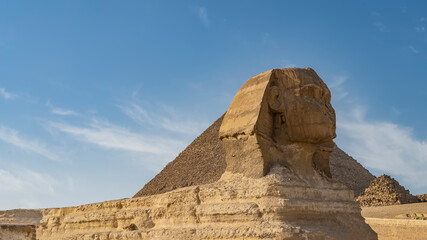 Fototapeta na wymiar The mysterious sculpture of the Great Sphinx is carved out of stone. Profile view. In the background is an ancient pyramid, blue sky with clouds. Egypt. Giza