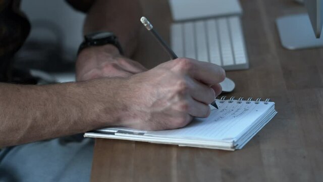 Close up side view of young man writing down notes with pen on paper notepad. Right handed businessman writing daily tasks in organizer.Guy sitting at table in front of computer making to do list. 
