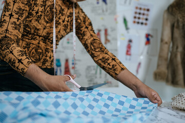 Cropped image of Asian young male tailor cutting piece of cloth at table in fashion studio. Designer and LGBTQ Concept.
