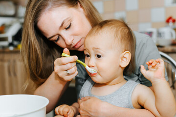 Mother with spoon feeds her baby puree while sitting in kitchen at home. Close-up child eating breakfast with mom. Motherhood, baby care, healthy food for children concept
