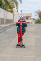 Cute little girl in winter clothes riding scooter on road