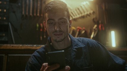 Young handsome blacksmith man sitting in front of the workbench, resting and using smartphone