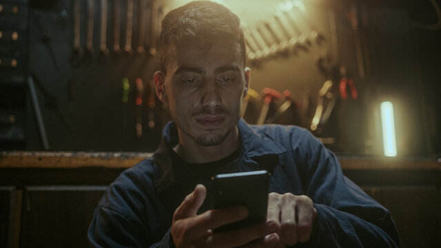 Young handsome blacksmith man sitting in front of the workbench, resting and using smartphone