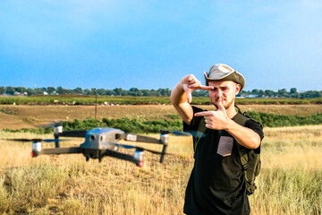 Naklejka na ściany i meble a young tourist guy in a hat shoots landscapes of the area on a quadcopter holding a frame made of fingers in front of him