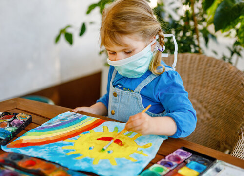 Little toddler girl in medical mask painting rainbow with water colors during pandemic coronavirus quarantine disease. Preschool child in nursery or kindergarten.