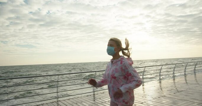 Young woman with long ponytail in mask jacket and leggings runs along wet boardwalk against stormy sea waves in windy weather slow motion