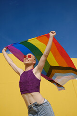 Low angle view of a young happy non-binary person holding and waving a rainbow flag over head.