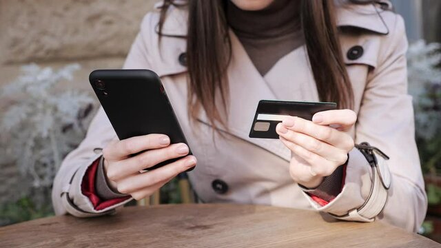 Close up female hands woman holding bank card, entering cvv code or payment information in mobile internet shopping application outdoors in the terrace of the restaurant, services online, purchase.
