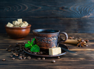 Hot, fragrant black coffee in a dark brown clay cup on a dark wooden table. Coffee beans and mint leaves on a saucer. Pieces of cane sugar in a clay bowl in the background