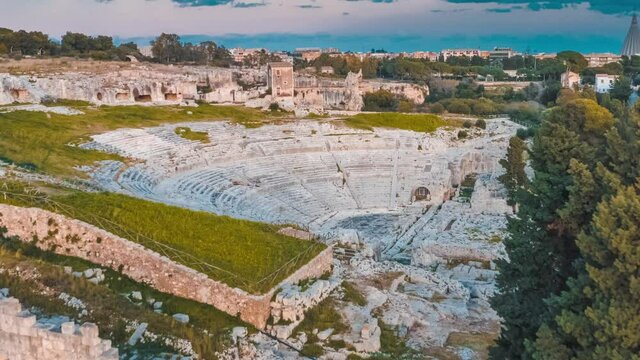 Stunning aerial view of the ancient Greek Theatre of Syracuse in Sicily, Italy. The historic stone ruins of the amphitheater are surrounded by green nature and trees.