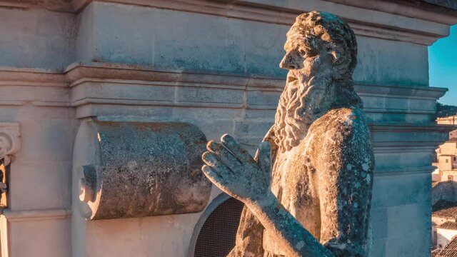 A close-up shot of an ancient stone statue of a praying saint on a historic building facade. The weathered sculpture is beautifully illuminated by warm evening sunlight.