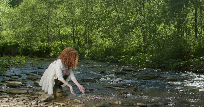 Side view on redhead curly caucasian female touching water, splashing it, have rest sitting near lake in forest, female in white beautiful dress enjoy nature. alone. in the evening.