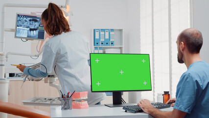 Assistant using horizontal green screen on computer while dentist preparing equipment for oral care examination in dental cabinet. Man looking at chroma key and mockup template on monitor