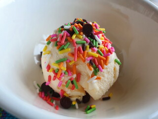 Homemade ice cream one scoop of vanilla is placed in a white cup and sprinkled with rainbow jimmies and chocolate rounds.
