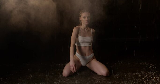 Beautiful leggy and booty athletic fitness model, wearing white underwear, with wet skin, sit on floor under water drops in theatrical smoke on black background. aqua studio photoshoot