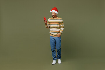 Full body young african man in knitted sweater red Christmas Santa Claus hat hold in hand use mobile cell phone isolated on plain green khaki background studio Happy New Year 2022 celebration concept
