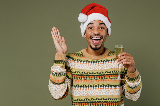 Young african man in knitted sweater red Christmas Santa Claus hat hold wineglass drink champagne say cheers isolated on plain green khaki background studio. Happy New Year 2022 celebration concept. - Powered by Adobe