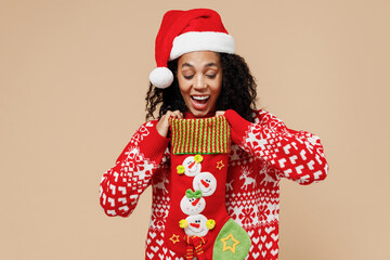 Happy young african american Santa woman in Christmas hat sweater hold decorative stocking isolated on pastel beige background studio portrait. Happy New Year celebration merry x-mas holiday concept.