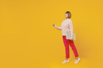 Full body elderly fun woman 50s wears pink casual knitted sweater hold in hand closed laptop pc computer walking go point finger aside on workspace isolated on plain yellow background studio portrait