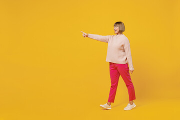 Fototapeta na wymiar Full body side view elderly smiling happy fun amazed woman 50s with bob haircut wear pink casual knitted sweater walk go point index finger aside isolated on plain yellow background studio portrait