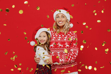 Young smiling woman in sweater hat have fun with child baby girl 6-7 years old. Mommy little kid daughter toss up confetti isolated on plain red color background studio. New Year love family concept.