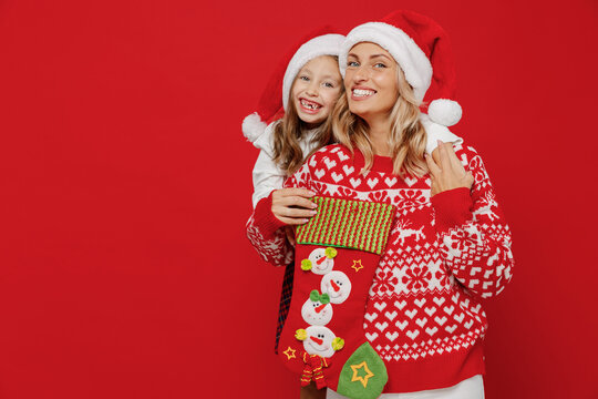 Young happy woman in sweater hat have fun with child baby girl 6-7 years old. Mommy little kid daughter look at gift sock stocking isolated on plain red background studio New Year love family concept.