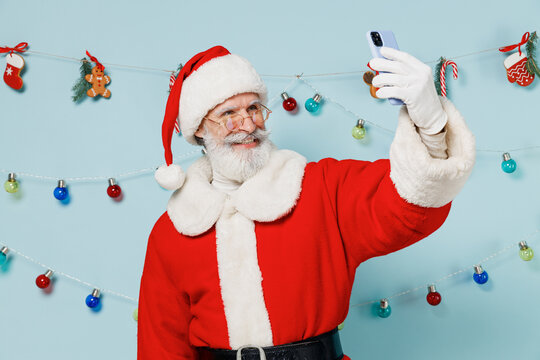Old bearded Santa Claus man 50s wears Christmas hat red suit doing selfie shot on mobile cell phone isolated on plain blue background studio. Happy New Year 2022 celebration merry ho x-mas concept