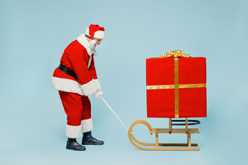 Full length sad old Santa Claus man in Christmas hat red suit clothes pull sled with gift box isolated on plain blue background studio. Happy New Year 2022 celebration merry ho x-mas holiday concept.